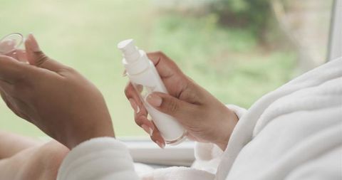 Sunlit closeup showing woman in white bathrobe dispensing moisturizer from pump bottle
