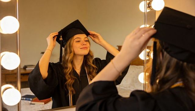 Graduate adjusting mortarboard at lighted vanity mirror wearing gown and honor cords