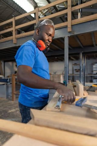 Mature african american craftsman measuring wood plank wearing goggles in workshop
