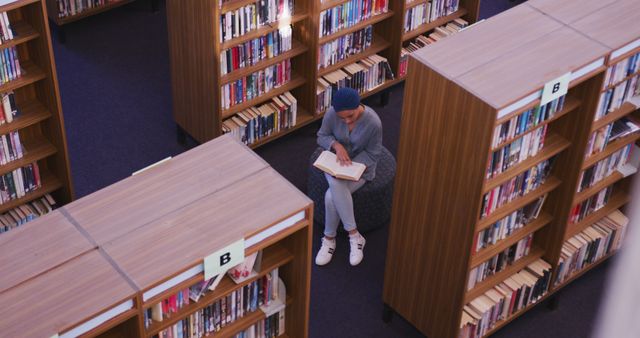 Woman engrossed in book sits alone in between tall bookshelves in library. Ideal for materials about education, libraries, reading habits, solitude, learning environments.