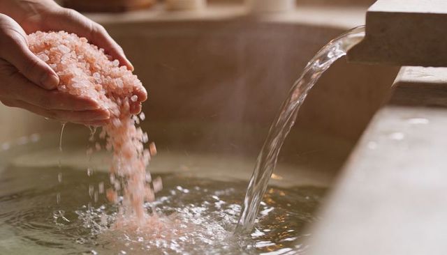 Hands Pouring Himalayan Pink Bath Salt into Stone Spa Basin with Flowing Water