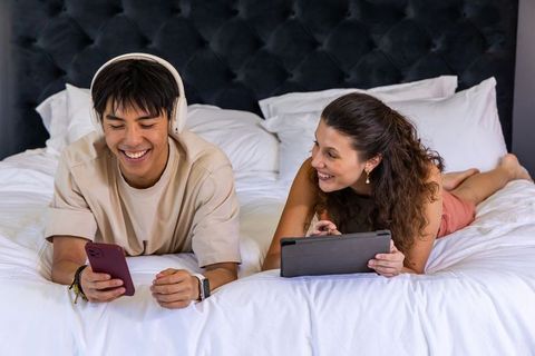 Multicultural couple relaxing on bed using smartphone and tablet with headphones and smartwatch