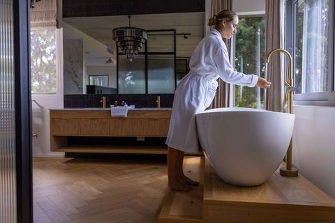 Woman in bathrobe reaching for gold faucet by freestanding tub in modern wooden bathroom