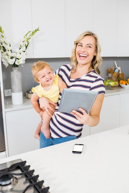 Smiling mother holding her baby girl while using a digital tablet in a modern kitchen. Ideal for themes related to family, parenting, technology in daily life, and modern home environments. Can be used in advertisements, blogs, and articles about family life, digital parenting, and home technology.