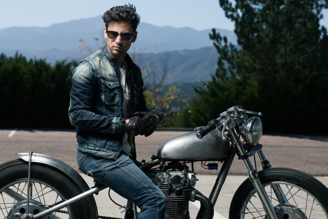Young man, sporting denim jacket and sunglasses, sitting confidently on classic motorcycle. Taken outdoors against scenic mountain backdrop, highlighting a mix of ruggedness and style. Use this image for promotions involving cool lifestyle, fashion, adventure or classic motorcycles.