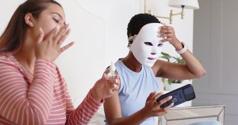 African american women applying serum and adjusting led face mask during skincare routine