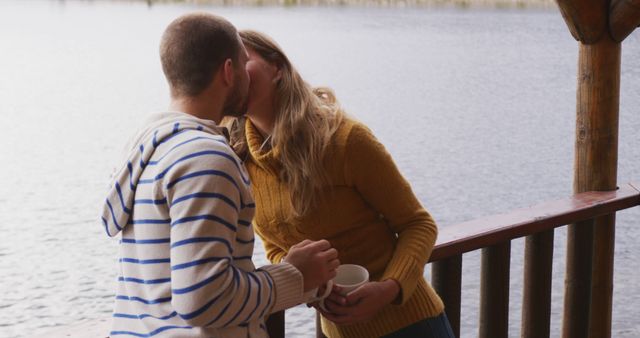 A couple is kissing on the porch of a lakeside cabin during an autumn day. They are dressed in cozy sweaters and are holding a mug. The scene is serene with calm water in the background. Great image for use in content about relationships, romantic getaways, autumn activities, and cozy lifestyles.