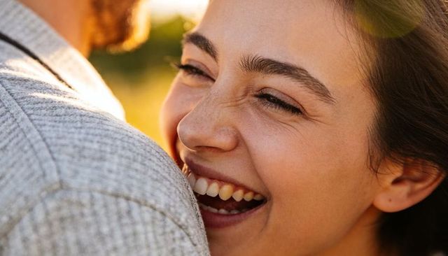 Laughing woman nestling into partner's shoulder at golden hour, candid sunlit close-up