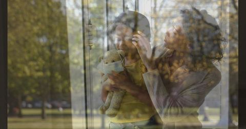 Mother comforting daughter at window holding masked teddy, park reflection outside