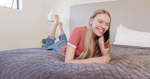 Young woman lying on bed smiling while talking on smartphone in bright modern bedroom