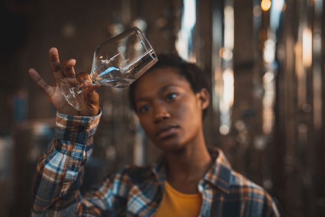 African american woman working at gin distillery, holding up a glass and inspecting product. work at an independent craft gin distillery business.