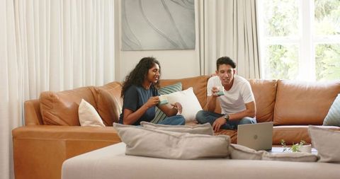 African american couple relaxing on tan leather sectional holding coffee cups with laptop