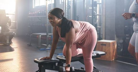 Black woman performing single-arm dumbbell row on bench with trainer in daylight gym