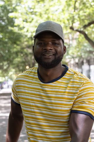 Smiling african man wearing gray cap and yellow striped tee on tree-lined urban walkway