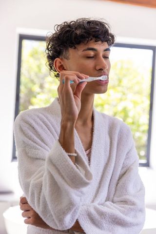 Person in bathrobe brushing teeth by sunlit window