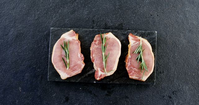 Three uncooked bacon steaks are garnished with fresh rosemary and placed on a slate stone background, making a visually appealing display of raw ingredients. Ideal for use in content related to cooking, culinary arts, food preparation, organic ingredients, recipes, or gourmet dining. It can also be useful for illustrating articles on meat, cuisine, kitchen techniques, or healthy eating.