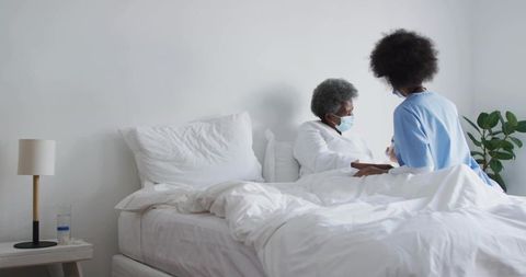 Nurse comforting senior patient at bedside wearing masks in bright hospital room