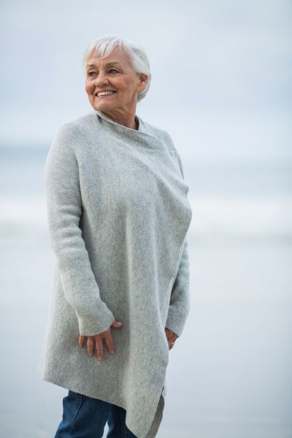 Elderly woman wearing a light grey sweater and standing on a beach. She is smiling and looks content. Ideal for use in articles promoting senior health, outdoor activities for older adults, and senior lifestyle content.