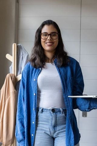 Young woman standing holding laptop wearing denim shirt and glasses in home workspace