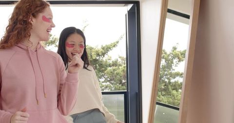 Friends applying pink undereye patches in mirror on balcony, morning self-care loungewear