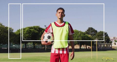 Young soccer player standing on grass field holding ball wearing neon bib and striped kit