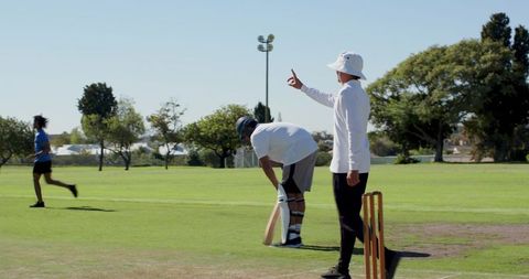 Cricket umpire signaling out raising finger on sunny park pitch with batsman and stumps