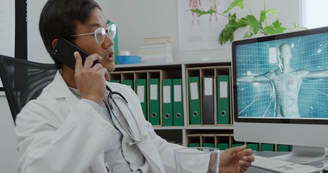 Asian man in a lab coat is analyzing a 3D human model on a computer at the office. He's engaged in a phone conversation, discussing medical findings.