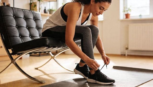 Woman tying running shoes at home gym corner sitting on modern chair preparing workout