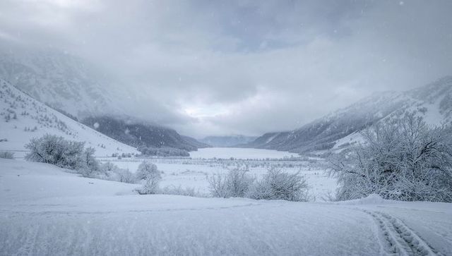 Snow-covered remote alpine valley with frozen lake and tracks under overcast snowfall