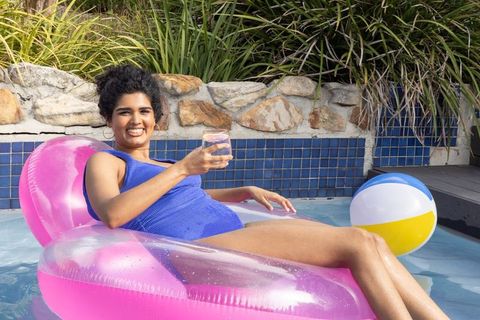 Indian woman relaxing on pink pool float in backyard holding refreshing drink