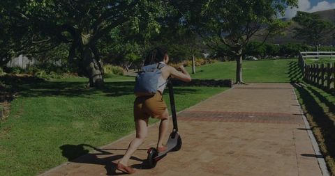 Young woman pushing electric scooter along sunlit brick park path wearing backpack