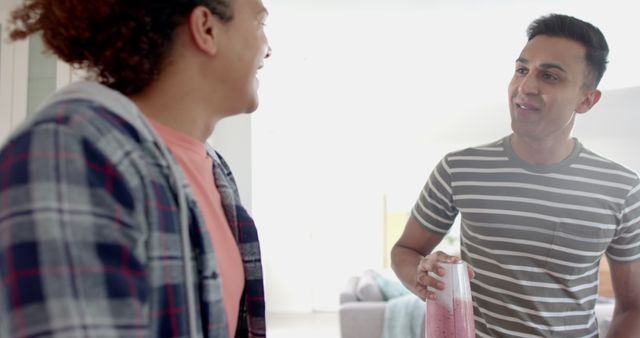 Two friends enjoying a conversation inside while holding a healthy smoothie drink. They appear relaxed and happy, suggesting a comfortable and friendly environment. Ideal for concepts related to friendship, healthy lifestyle, casual living, and home life.