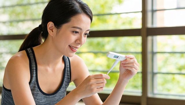 Smiling woman checking digital thermometer near sunlit window for wellness and self care