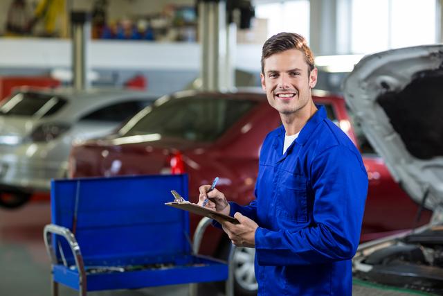 Smiling mechanic wearing blue overalls writes a checklist in an auto repair shop. Cars are being repaired in the background. Useful for themes related to automotive service, car maintenance, professional repair services, and mechanic training.