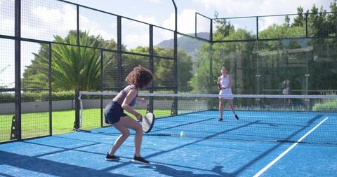 Two women playing padel on bright blue outdoor court hitting ball near net fitness match