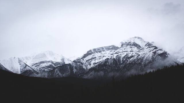 This striking image captures a range of snow-capped mountains under an overcast sky. Fog and mist swirl around the rugged peaks, creating a dramatic and cold atmosphere. Makes for a perfect background, wallpaper, or inspirational imagery in topics related to adventure, nature conservation, winter sports, or atmospheric scenery.