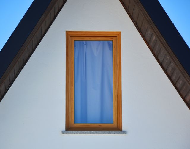 Minimalist design features prominent triangular roof structure with wooden framed window and blue curtain. Ideal for use in articles or advertisements exploring modern architecture, home design, or geometric simplicity.