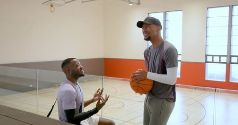 Two teammates laughing and bonding on bleachers with basketball in indoor gym for teamwork
