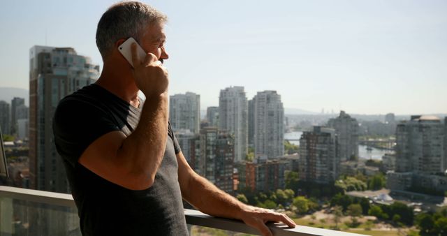 A middle-aged Caucasian man is engaged in a phone conversation on a balcony overlooking a cityscape, with copy space. His casual attire and relaxed posture suggest a personal call rather than professional.