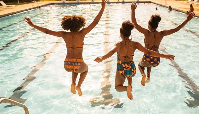 Three teenagers jumping into sunlit outdoor pool wearing colorful patterned swimsuits
