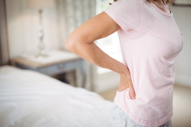 Senior woman standing in bedroom, holding lower back in discomfort. Useful for articles on elderly health, back pain relief, aging, and home care solutions.