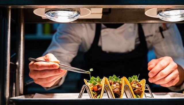 Chef plating gourmet mini tacos with microgreens and drizzle at kitchen pass