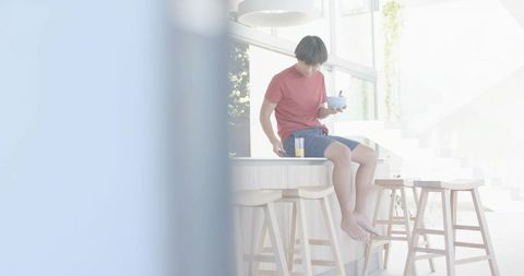 Young Asian man sitting on kitchen island holding bowl and juice in bright modern kitchen