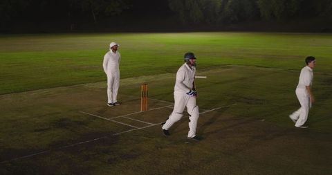 Night cricket match under floodlights with batsman running between wickets in white kit