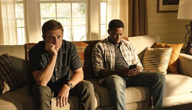 Two men sitting on sofa, pensive white man and black man scrolling smartphone in cozy living room