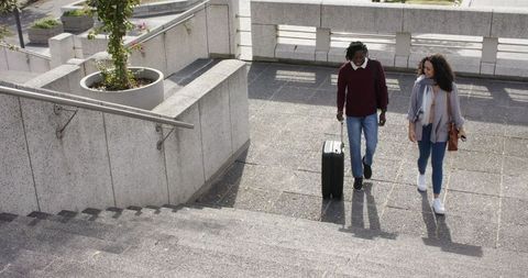 Diverse young couple walking sunlit urban plaza pulling suitcase and carrying bag