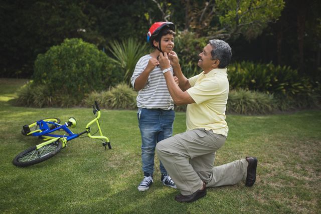 Grandfather kneeling on grass helping grandson with bicycle helmet in park. Bicycle lying on ground nearby. Perfect for themes of family bonding, outdoor activities, childhood safety, and intergenerational relationships.