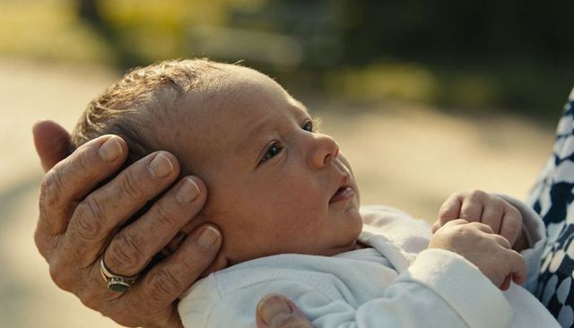 Elderly hands cradling newborn in white onesie, ornate ring and tender gaze in warm light
