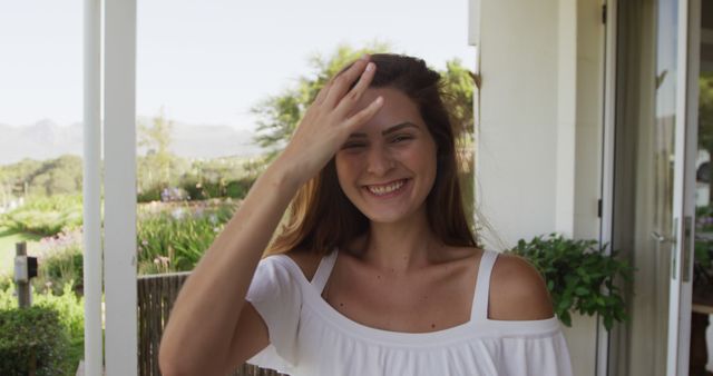 Young woman enjoying a sunny day outdoors, standing on a porch with greenery in the background. Perfect for use in advertisements, lifestyle blogs, social media posts, and websites promoting outdoor activities, fashion, and positive moments.