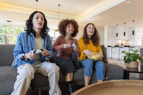 Multicultural friends playing video games on sofa eating chips in modern cozy living room